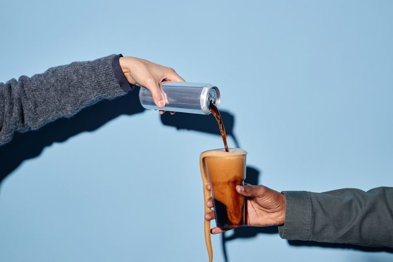 A hand is seen pouring canned coffee into a glass.